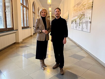 Zwei Personen stehen in einem langen Flur mit großen Fenstern und Bildern an der Wand.