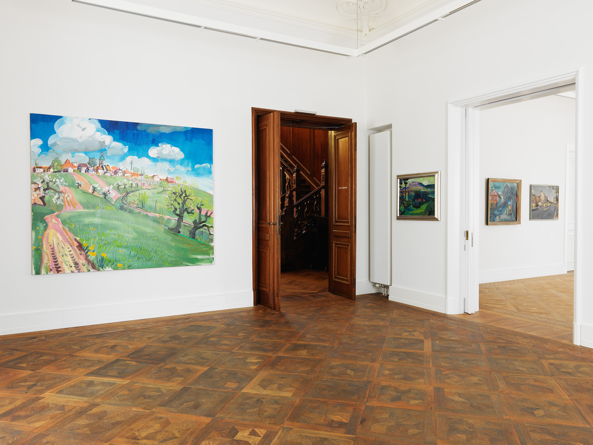 Ein Ausstellungsraum in der Kunstvilla mit Gemälden an der Wand. Durch die beiden Eingänge führen einmal in das Treppenhaus und dann in einen weiteren Ausstellungsraum.