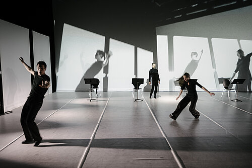 Personen in schwarz gekleidet auf der Bühne Hinter ihnen spiralförmige Projektion der Stühle auf der Bühne