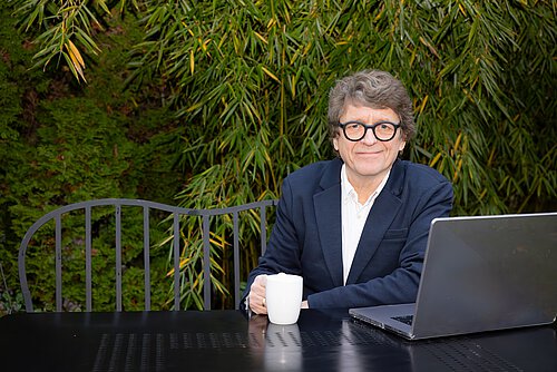 Älterer Mann mit grauen Haaren sitzt an einem schwarzen Metalltisch im Freien vor einem Laptop und hält eine weiße Tasse