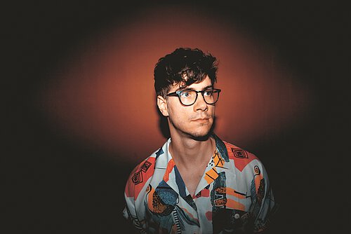 Junger Mann mit lockigem Haar trägt ein bunt gemustertes Hemd vor einem orangefarbenen Hintergrund.