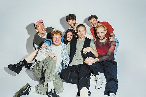 Sieben Personen sitzen auf dem Boden, der Hintergrund ist neutral weiß gehalten. Alle schauen mit einem Lächeln in die Kamera.