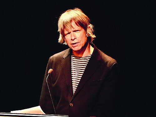 Mann mit halblangem Haar, ernst, schwarzer Blazer, vor schwarzem Hintergrund