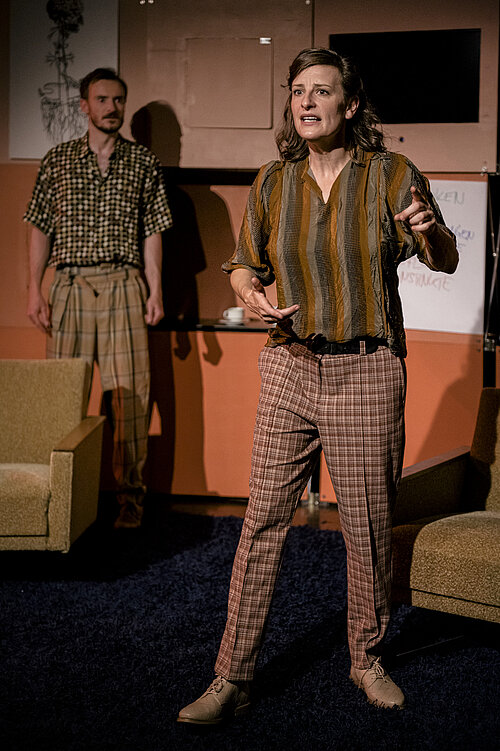 Person mit schulterlangen Haaren trägt gestreiftes Hemd und karierte Hose, steht gestikulierend in einem Raum mit braunen Möbeln und Wänden.