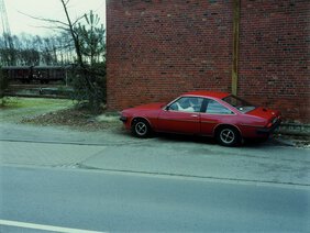 Aufnahme zeigt ein rotes Auto am Rande einer Straße stehend, eine Person ist hinter dem Steuer zu erkennen. Das Auto parkt vor einem Backsteinhaus
