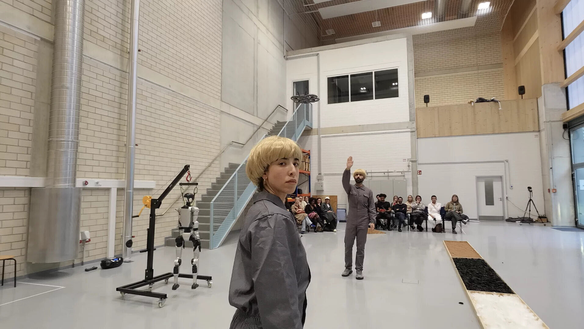 Person mit blonden kurzen Haaren in grauer Arbeitskleidung steht in einer großen Halle, im Hintergrund sitzt eine Gruppe von Menschen und eine weitere Person hebt die Hand