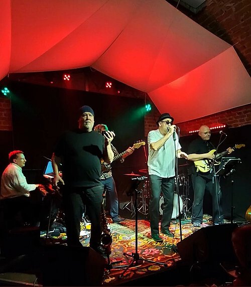 Auf einer Bühne stehen verschiedene Musiker, die bei rotem Hintergrundlicht performen.