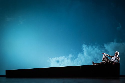 Ein Mann liegt auf einem dunklen Bühnenelement, im Hintergrund ist ein Himmel projiziert. 