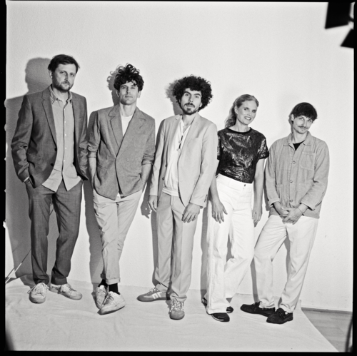 Fünf Personen stehen vor einer weißen Wand, alle tragen legere Kleidung mit Jacken und Hosen, zwei Personen haben lockiges Haar, eine Person trägt ein glänzendes Oberteil.