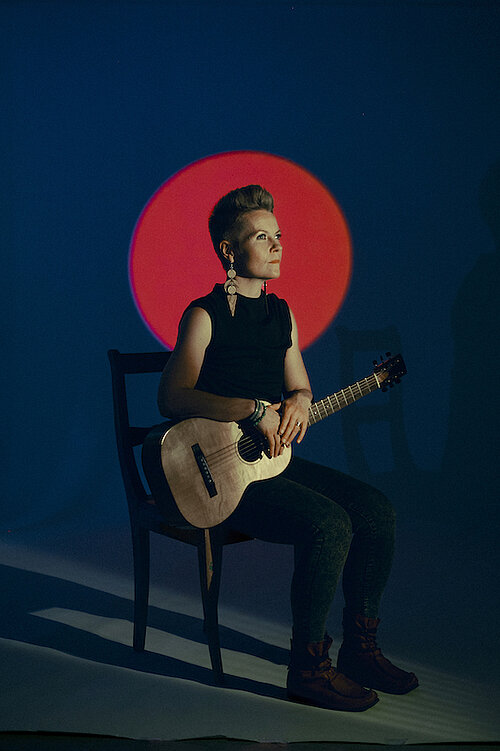 Person sitzt auf einem Stuhl und hält eine Gitarre, im Hintergrund ein roter Kreis vor dunkelblauem Hintergrund.