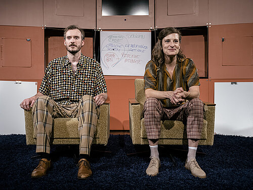 Zwei Personen sitzen auf beigen Sesseln, tragen karierte Hemden und Hosen mit verschiedenen Mustern. Im Hintergrund steht ein Flipchart mit handschriftlichen Notizen.