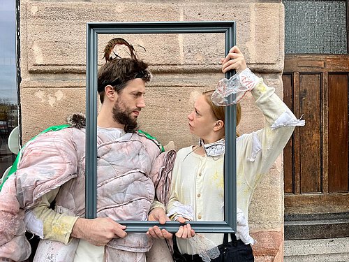 ein Mann in Käferkostüm und eine Frau in weißer Bluse rahmen ihre Gesichter mit einem leeren Bilderrahmen