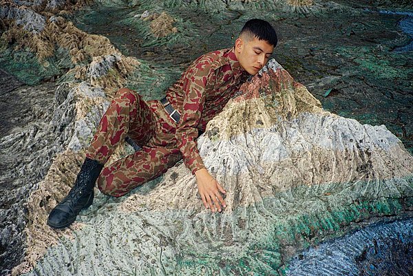 Ein Mann in Camouflagekleidung schmiegt sich an einen Fels inmitten einer steinigen Landschaft. Der Fels nimmt teilweise die Farben der Camouflage auf