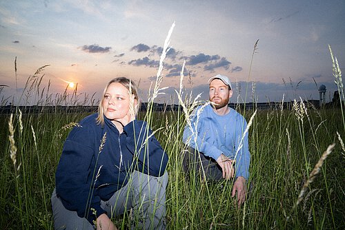 Ein Mann und eine Frau sitzen in einem Wiesenfeld, die Sonne geht langsam im Hintergrund unter