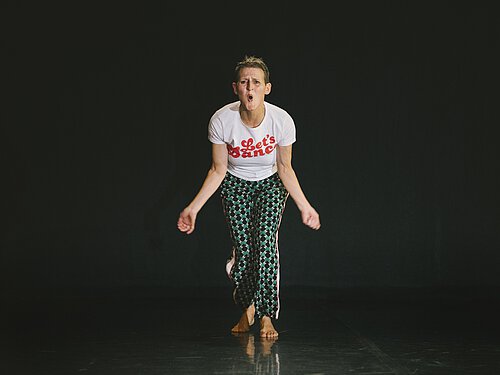 Frau mit kurzem Haar in weißem T-Shirt mit rotem Schriftzug 'Let's Dance' und gemusterten Hosen tanzt barfuß vor schwarzem Hintergrund