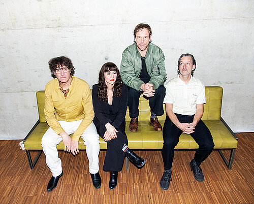 Vier Personen sitzen auf einem gelben Sofa vor einer Betonwand, alle mit verschränkten Händen oder Armen.