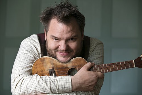 Mann lehnt sich mit Kinn auf Ukulele und blickt in die Kamera