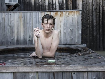 Junger Mann lehnt am Beckenrand eines Ausßenwhirlpools mit Holzverkleidung, raucht eine Zigarette und blickt in die Kamera.