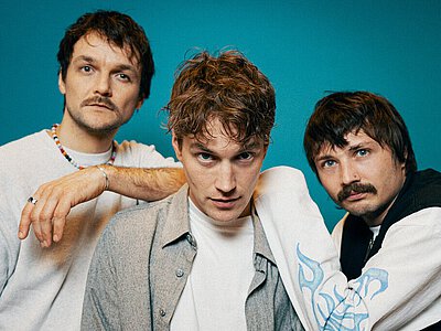 Drei junge Männer stehen vor blauem Hintergrund, einer trägt ein weißes T-Shirt mit bunter Halskette, ein anderer ein graues Hemd über weißem T-Shirt, der dritte ein weißes Shirt mit schwarzer Weste.