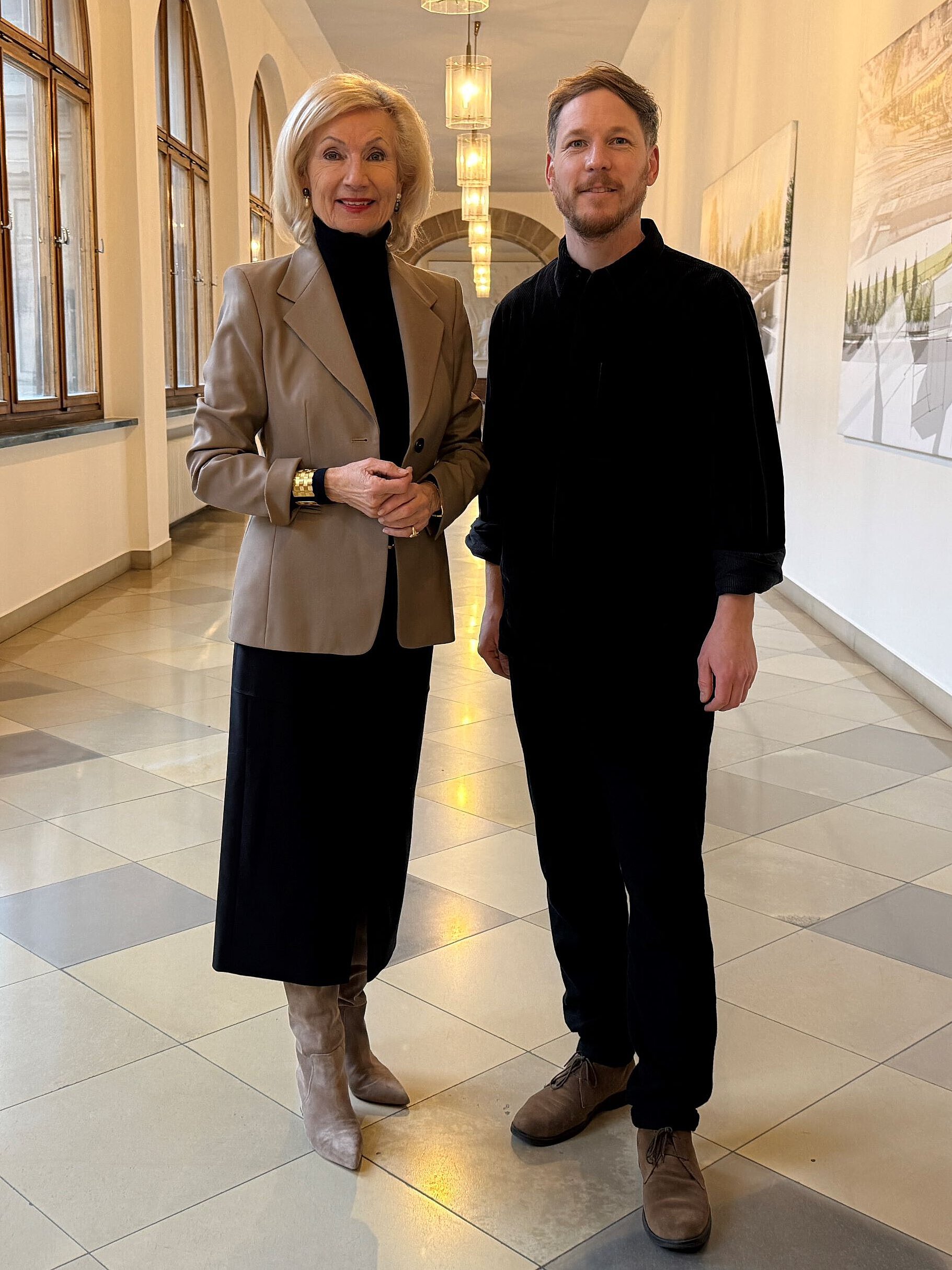 Zwei Personen stehen in einem langen Flur mit großen Fenstern und Bildern an der Wand.