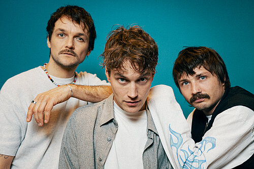 Drei Männer in lässiger Kleidung stehen vor einem türkisem Hintergrund