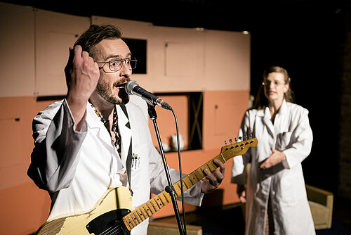 Person in weißem Kittel spielt E-Gitarre vor Mikrofon, weitere Person in weißem Kittel im Hintergrund mit verschränkten Armen.