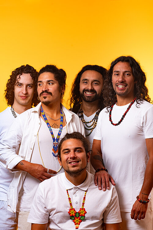 Gruppenfoto der Band vor einem gelben Hintergrund, sie tragen weiße Poloshirts und manche von ihnen bunte Ketten