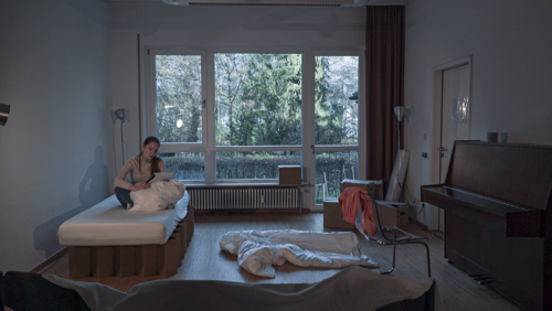 Eine Frau sitz auf einem Bett und liest, ihr Zimmer ist unordentlich.