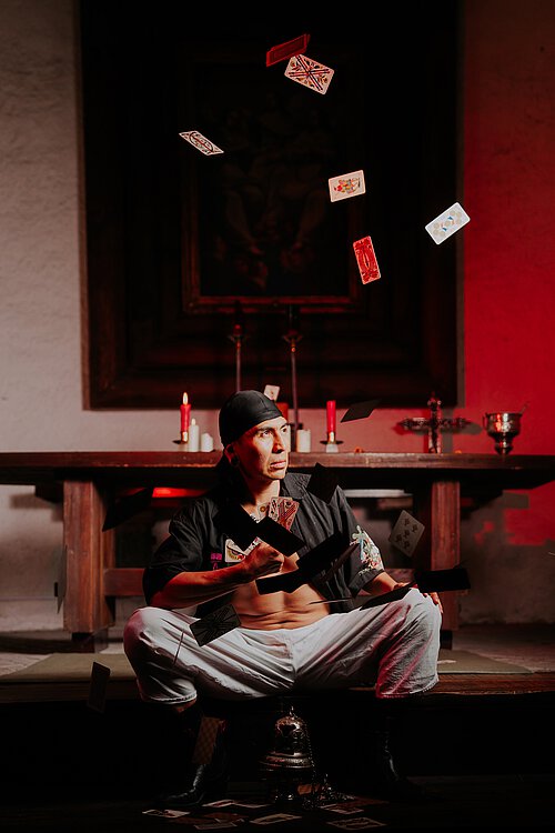 Mann mit schwarzer Kopfbedeckung sitzend, um ihn herum fliegende Karten