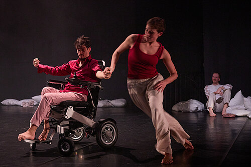 Zwei Tänzer auf einer Bühne, einer im Rollstuhl hält die Hand des anderen, eine dritte Person sitzt im Hintergrund auf dem Boden neben Kissen.