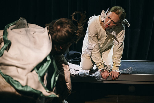 Mann in Käferkostüm kniend von hinten und Frau in weißem Overall kniend schauen sich an