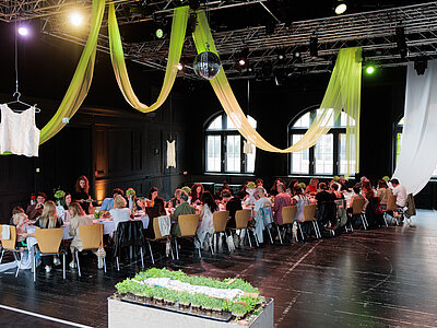 Eine lange Tafel, an dem viele Menschen sitzen steht im Festsaal des Künstlerhauses.