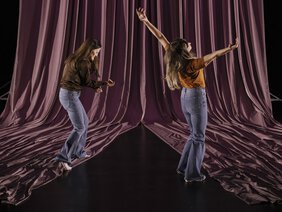Zwei kanghaarige Frauen in Jeans gestikulierend auf Bühne vor pinkfarbenen Vorhängen