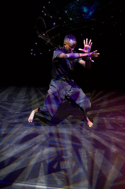 Tanzender Mann in schwarzem Oberteil und grauen Hosen, kniet auf dem Boden mit erhobenen Händen. Geometrische Muster und Lichtstrahlen auf dem Boden.