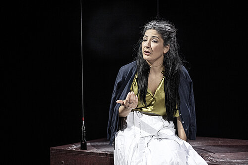 Eine Person mit langen, schwarzen Haaren trägt ein gelbes Oberteil und einen weißen Rock. Sie sitzt auf einer Plattform und gestikuliert mit der Hand.