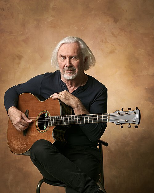 Älterer Mensch mit schulterlangem weißen Haar sitzt auf Stuhl und hält akustische Gitarre im Schoß, trägt schwarzes Langarmshirt und schwarze Hose.