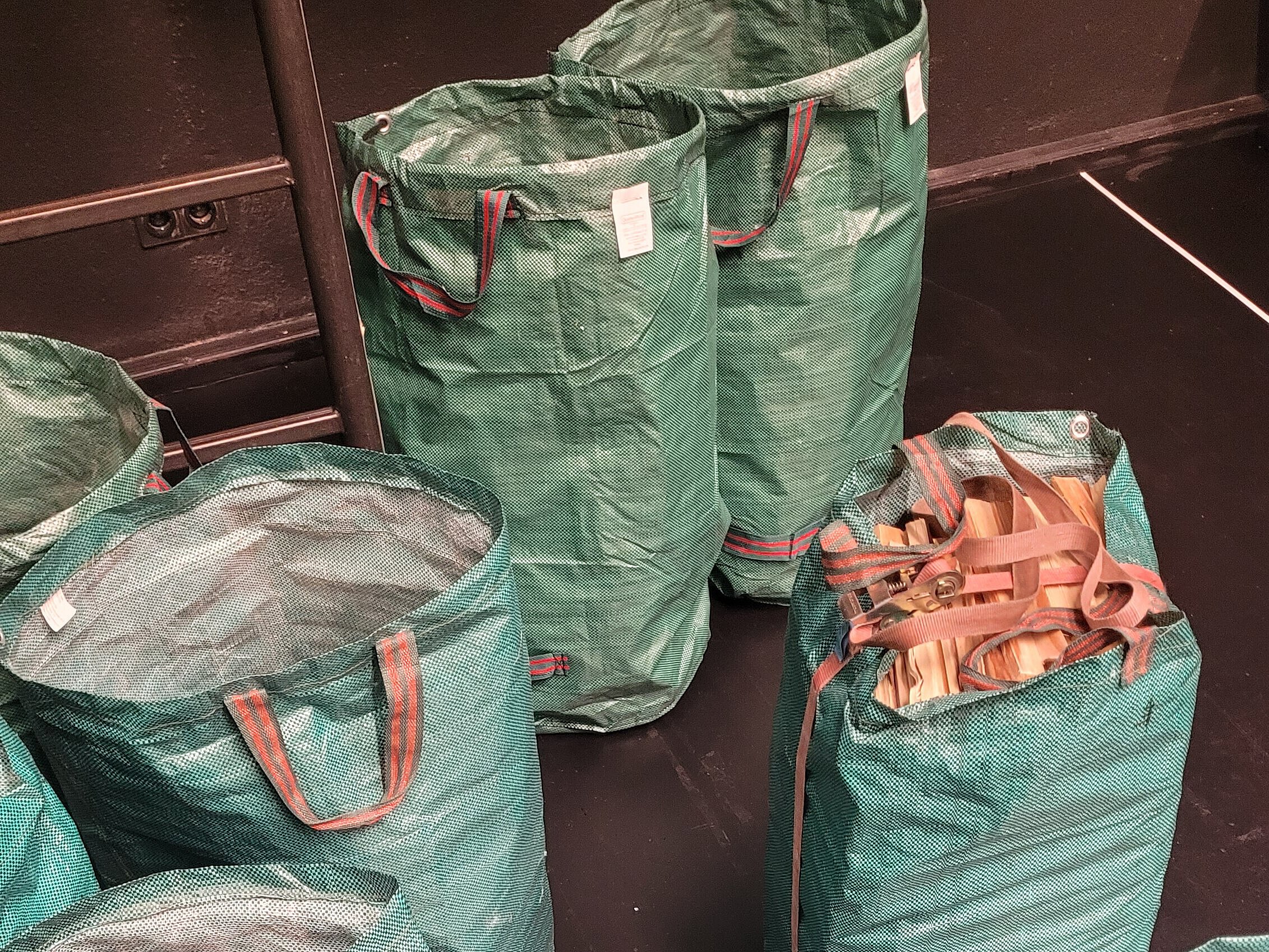 Grüne, robuste Säcke mit roten Griffen, einige gefüllt mit Holzscheiten, auf dunklem Boden verteilt