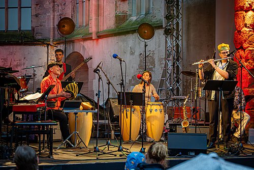 Auf einer Bühne spielen vier Musiker*innen: Zwei sind an Trommeln, einer an der Gitarre und einer an der Querflöte. 