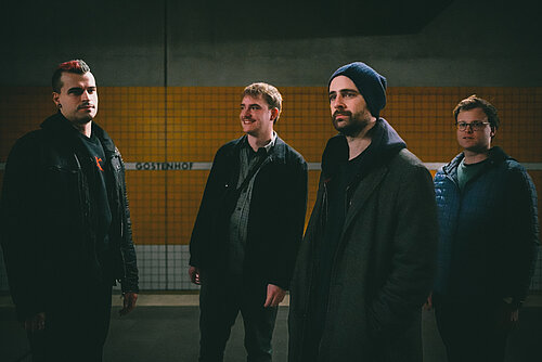 Vier dunkel gekleidete Männer stehen an einem unterirdischen Bahngleis. 