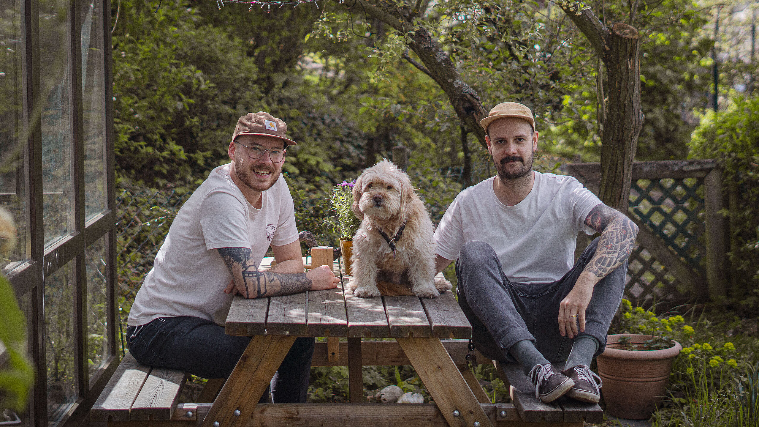 Zwei Männer mit Tattoos und weißen T-Shirts sitzen an einem Holztisch im Garten, zwischen ihnen sitzt ein kleiner Hund