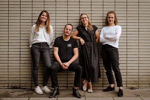 Vier Personen vor einer Wand mit horizontalen Linien, drei stehen, eine sitzt, alle tragen schwarze oder weiße Kleidung.