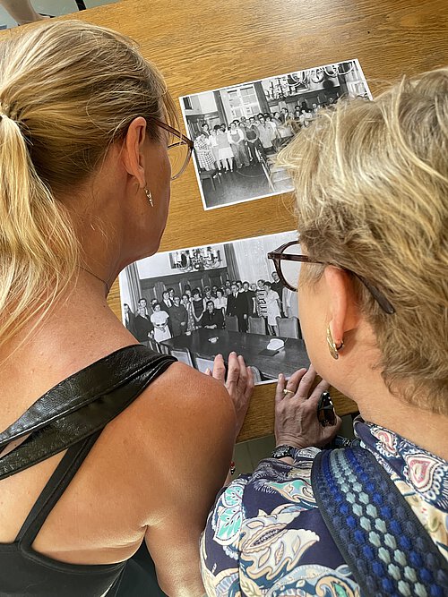 Zwei Frauen mit Brille und Ohrringen betrachten schwarz-weiße Gruppenfotos auf einem Holztisch.
