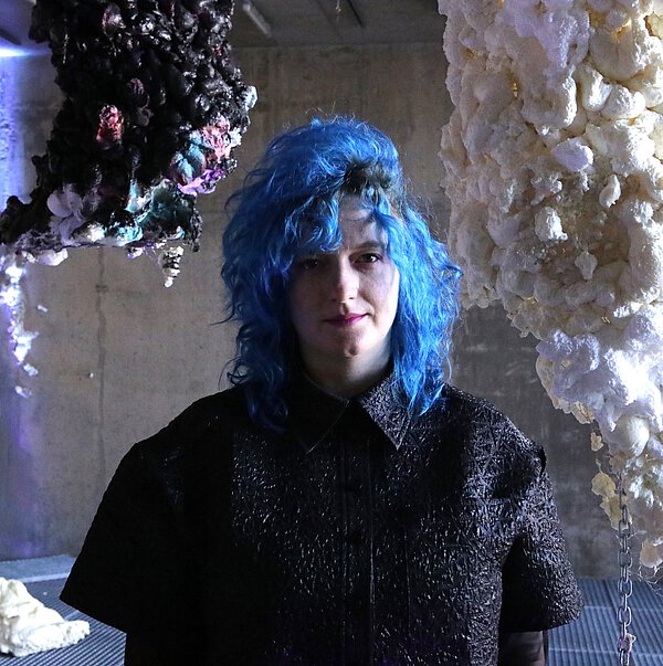 Vor verschiedenen Skulpturen steht eine Frau mit blauen Haaren, die ein schwarzes Hemd trägt