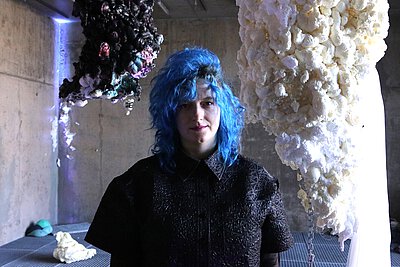 Vor verschiedenen Skulpturen steht eine Frau mit blauen Haare, die ein schwarzes Hemd trägt