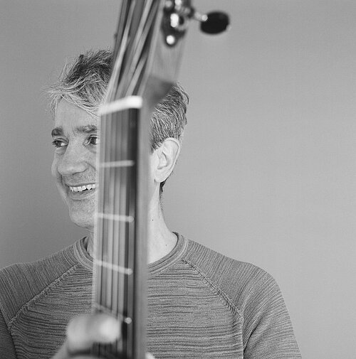 Schwarz-weiß-Porträt eines Mannes hinter einem aufrecht gehaltenen Bass mit sichtbarem Hals und Kopf des Instruments