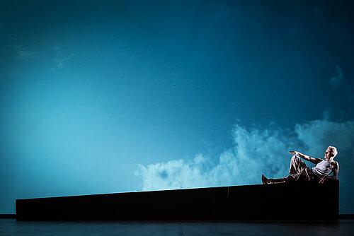 ein Mann liegt seitlich auf einer schrägen Erhöhung, im Hintergrund blauer Himmel mit wenig Wolken projiziert