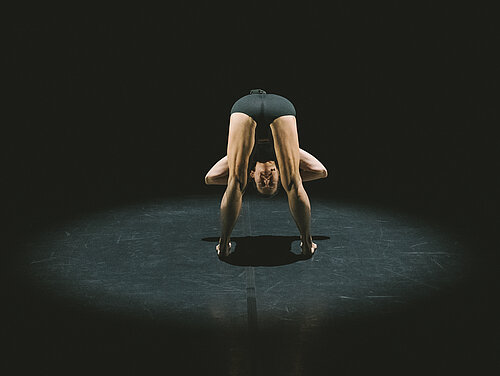 Person in schwarzer Sportkleidung steht gebeugt, Hände auf dem Boden, Kopf zwischen den Beinen, auf dunklem Boden mit Lichtkreis