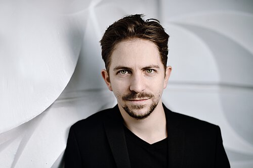 Person mit dunklem Sakko und schwarzem T-Shirt vor einer weißen, wellenförmigen Wand.