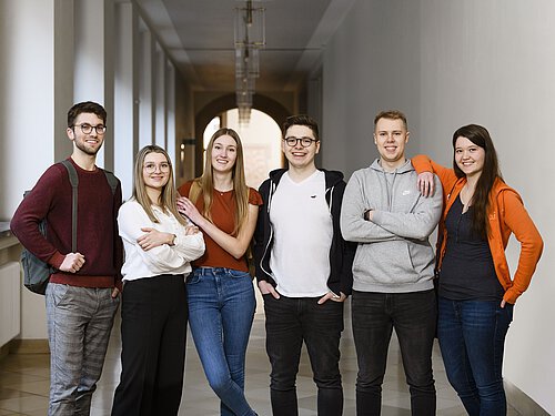 6 junge Menschen stehen in einem öffentlichen Gebäude nebeneinander und blicken freundlich und erwartungsvoll in die Kamera.