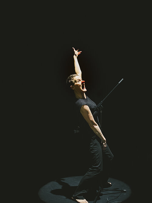 Frau in schwarzer Kleidung mit ausgestrecktem Arm und Golfclub in der Hand vor schwarzem Hintergrund.